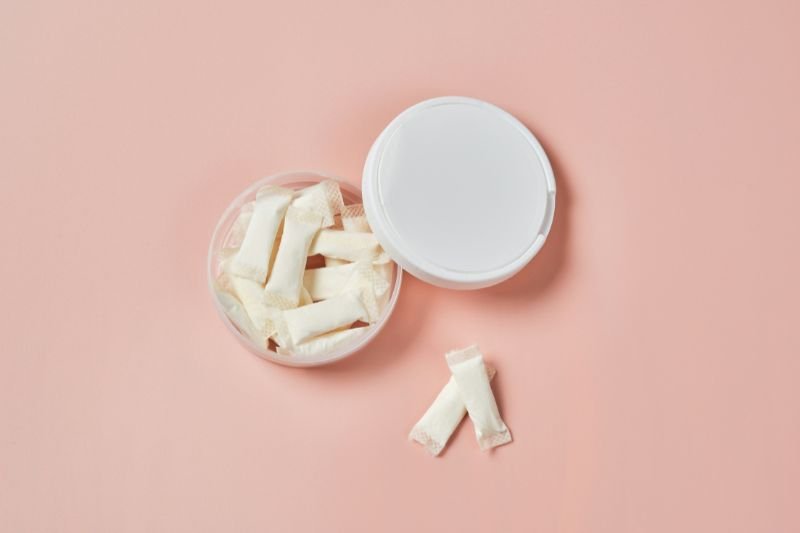 Open container of nicotine pouches with lid partially removed and two white pouches displayed on table surface